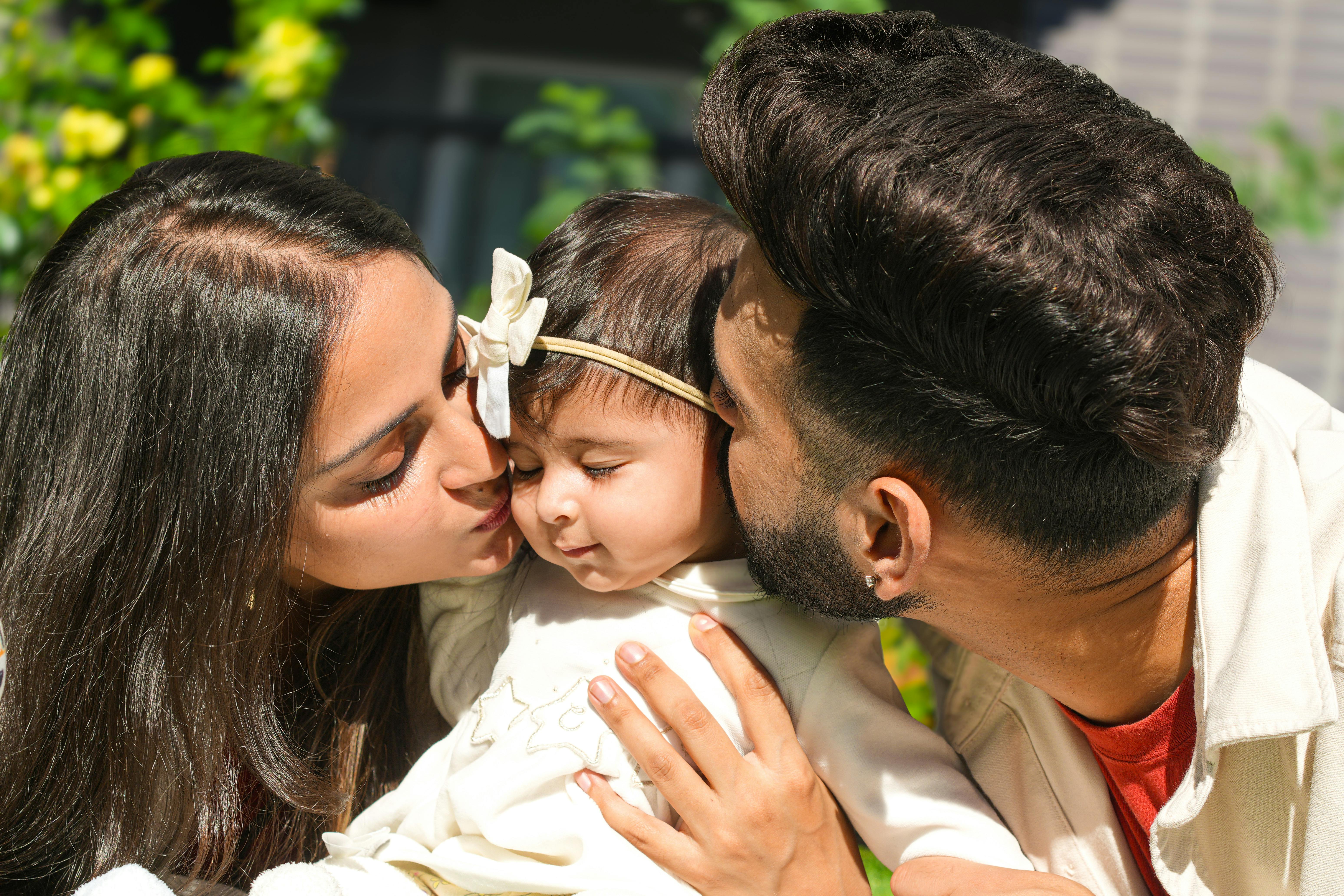 Tender moment of parents kissing their baby outdoors in bright daylight, showcasing family love.